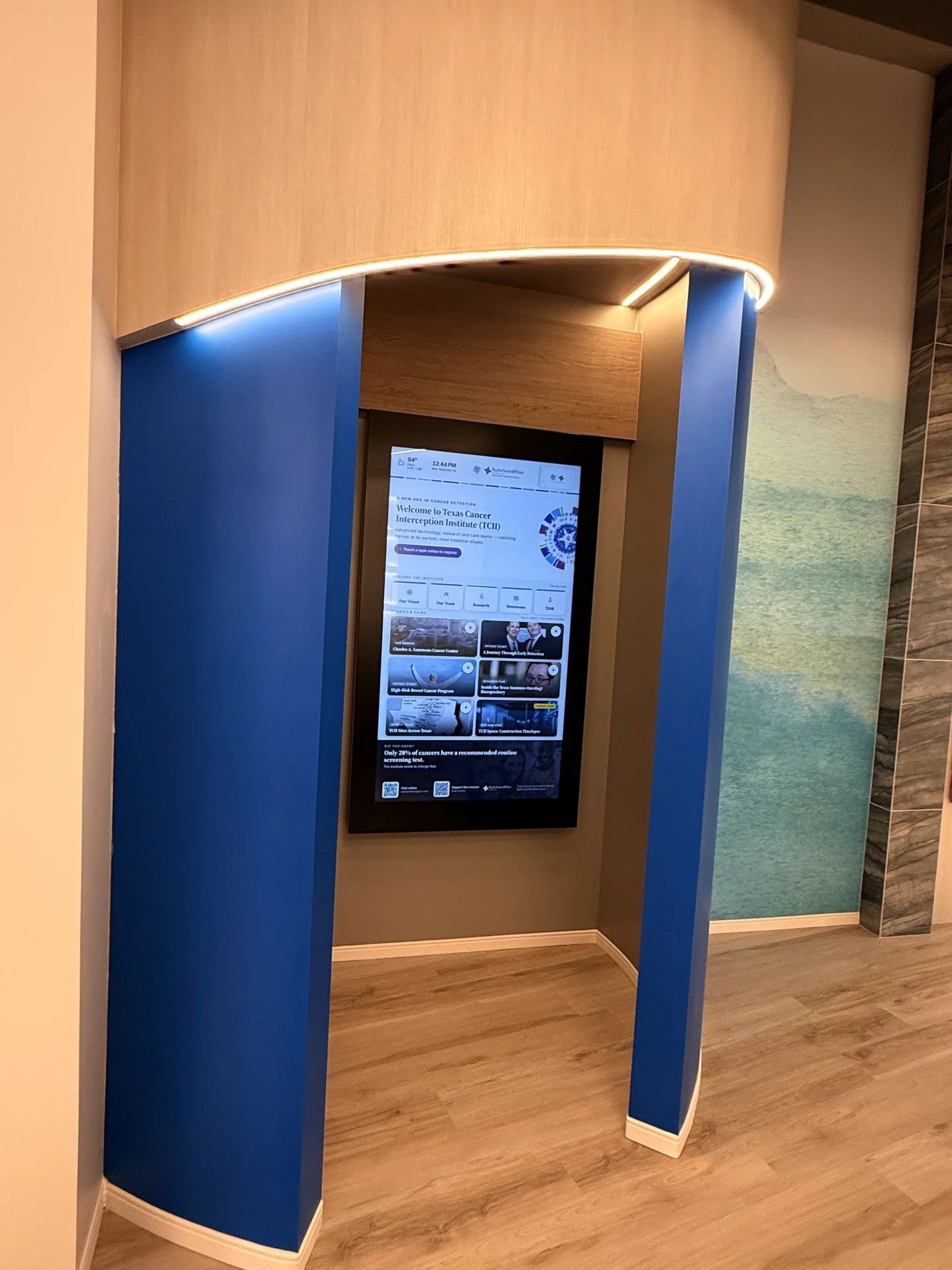 The installed portrait touchscreen in the TCII patient-story area, set into a blue architectural surround.